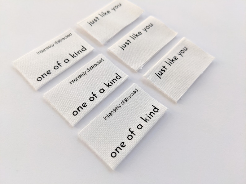 K&ouml;nnte beinhalten: Sechs wei&szlig;e Stoff-Etiketten mit schwarzem Text. Die Etiketten lauten "Intensely distracted one of a kind" und "Just like you".