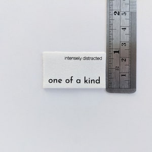 K&ouml;nnte beinhalten: Wei&szlig;es Stofflabel mit schwarzem Text, der "intensely distracted" und "one of a kind" lautet. Das Etikett wird an einem Lineal gemessen.