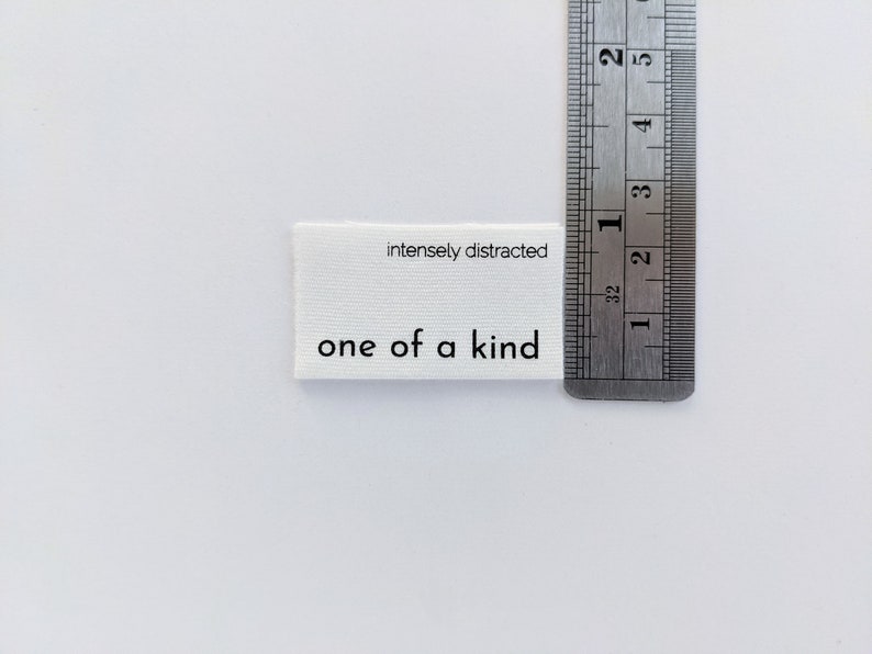 K&ouml;nnte beinhalten: Wei&szlig;es Stofflabel mit schwarzem Text, der "intensely distracted" und "one of a kind" lautet. Das Etikett wird an einem Lineal gemessen.