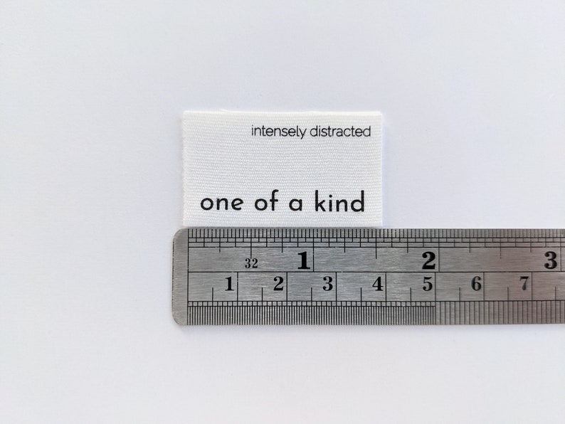 K&ouml;nnte beinhalten: Wei&szlig;es Stofflabel mit schwarzem Text, der "intensely distracted" und "one of a kind" lautet. Das Etikett wird mit einem Lineal gemessen.