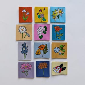 以下が含まれることがあります： 正方形の布の背景にそれぞれ刺繍された12個の花柄パッチのコレクション。パッチには、デイジー、バラ、ポピーなど、さまざまな花が鮮やかな色で描かれています。背景はオレンジ、イエロー、ピンク、ブルー、パープルの色合いです。