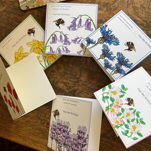 以下が含まれることがあります： 花とミツバチのイラストが描かれたグリーティングカードのコレクション。各カードには、スイセン、ブルーベル、ヤグルマギク、ラベンダー、バラなど、さまざまな花が描かれています。「Hap-Bee Birthday」の文字が各カードに印刷されています。