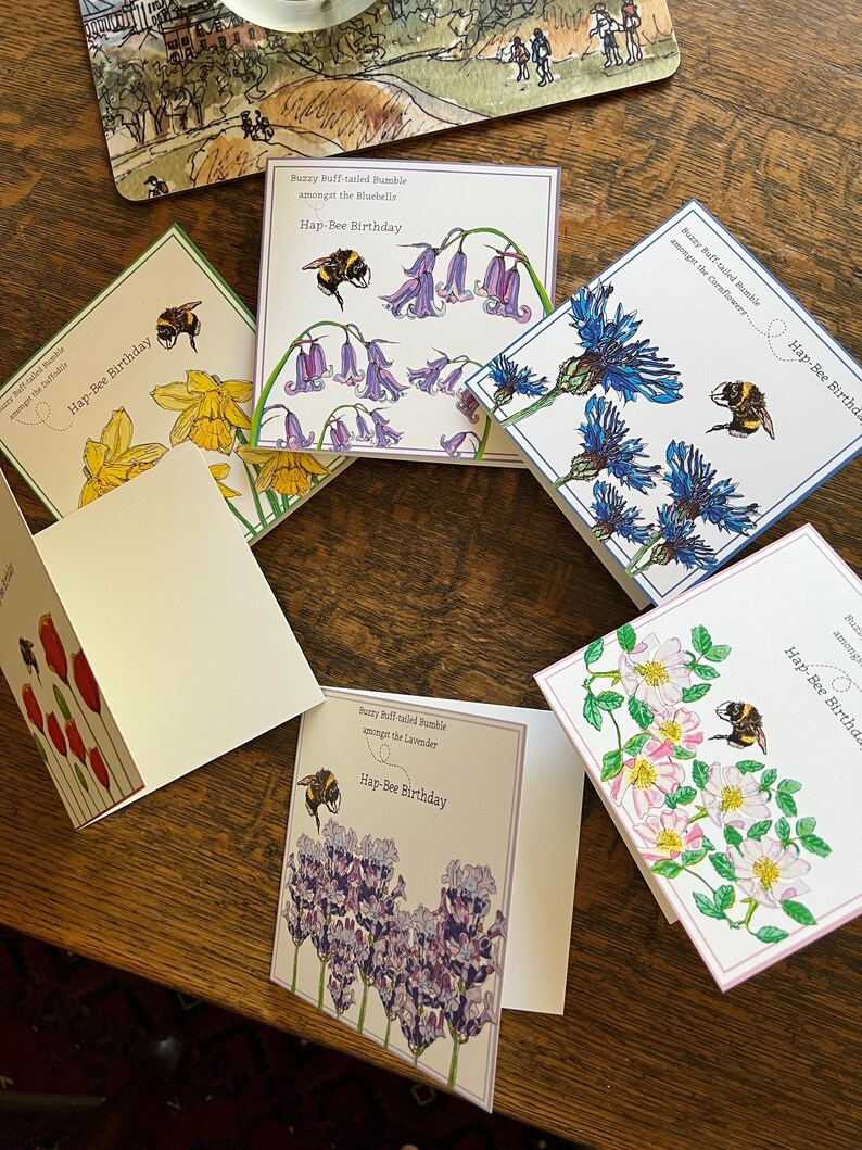 以下が含まれることがあります： 花とミツバチのイラストが描かれたグリーティングカードのコレクション。各カードには、スイセン、ブルーベル、ヤグルマギク、ラベンダー、バラなど、さまざまな花が描かれています。「Hap-Bee Birthday」の文字が各カードに印刷されています。