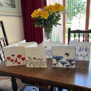 以下が含まれることがあります： 木製のテーブルに並べられた、花と蜂のイラストが描かれた「Hap-bee Birthday」グリーティングカードのコレクション。カードには、ポピー、バラ、ヤグルマギクなどの花が描かれています。黄色のバラの花瓶も近くにあります。