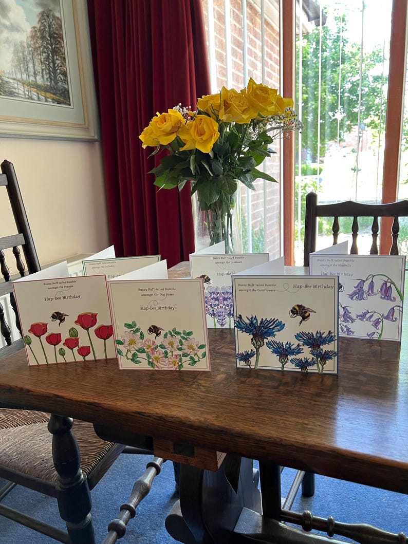 以下が含まれることがあります： 木製のテーブルに並べられた、花と蜂のイラストが描かれた「Hap-bee Birthday」グリーティングカードのコレクション。カードには、ポピー、バラ、ヤグルマギクなどの花が描かれています。黄色のバラの花瓶も近くにあります。