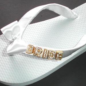 Può includere: Ciabatte bianche con un fiocco di raso e la parola "BRIDE" scritta in lettere dorate con strass.