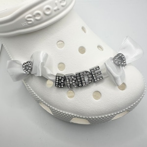 May include: White Crocs shoe with decorative embellishments. The word "BRIDE" is spelled out in silver letters with clear rhinestones. White satin bows with heart-shaped rhinestone accents are attached to the shoe.