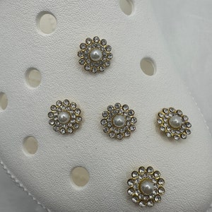 May include: Five gold-coloured charms with white pearl centres and clear rhinestones surrounding the pearls. The charms are on a white surface.