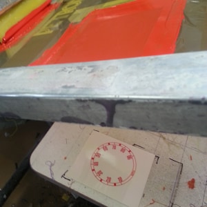 May include: A close-up of a screen printing process with a squeegee and orange ink being applied to a screen. A white paper with a red circle containing numbers from 10 to 80 is visible in the background.