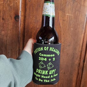 May include: Black and green neoprene can cooler with the text "Potion of Healing", "Common 2D4 + 2", "Drink Up! Might Need A Few To Do The Job."