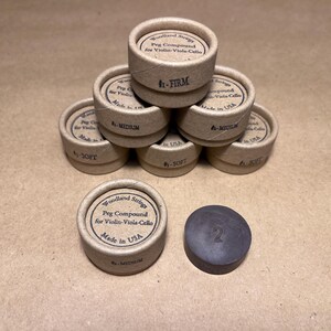 May include: A close-up of a stack of small cardboard containers labeled "Woodland Strings Peg Compound for Violin-Viola-Cello" with different firmness levels: "Firm", "Medium", and "Soft". There is also a single black peg compound in the image labeled "2".