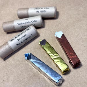 May include: Three small tubes of peg compound for violin, viola, and cello strings. The tubes are made of cardboard and have labels with the text "Woodland Strings Premium Peg Compound for Violin-Viola-Cello Made in USA". There are also three small bars of peg compound wrapped in foil, one silver, one gold, and one copper.