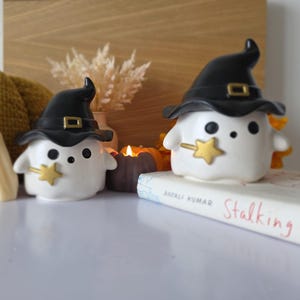 Peut inclure: Une composition sur le thème d'Halloween avec deux figurines de fantômes blancs portant des chapeaux de sorcière noirs avec des boucles dorées et des baguettes étoiles. Une bougie en forme de fantôme et une citrouille tricotée sont également présentes.