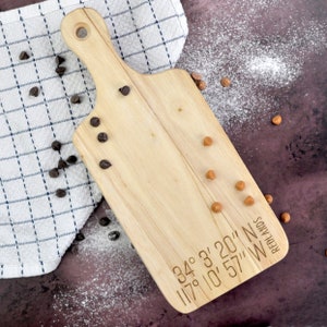 May include: A wooden chopping board with the coordinates "34° 3' 20" N 117° 10' 57" W" and the word "REDLANDS" engraved on the surface. The board is surrounded by brown chocolate chips and small, round, light brown sweets.