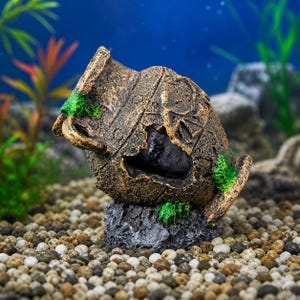 May include: A decorative aquarium ornament depicting a weathered, broken amphora. The brown and gold-toned ceramic pot is adorned with green moss and rests on a dark gray rock base. The background features aquatic plants and a blue backdrop, creating an underwater scene.