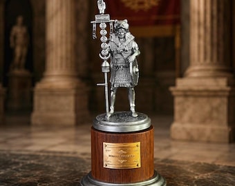 May include: A silver-colored Roman Centurion figurine on a wooden base with a metal plaque. The figurine is holding a spear and shield. The base is round with a silver trim. The background features columns and a red banner.