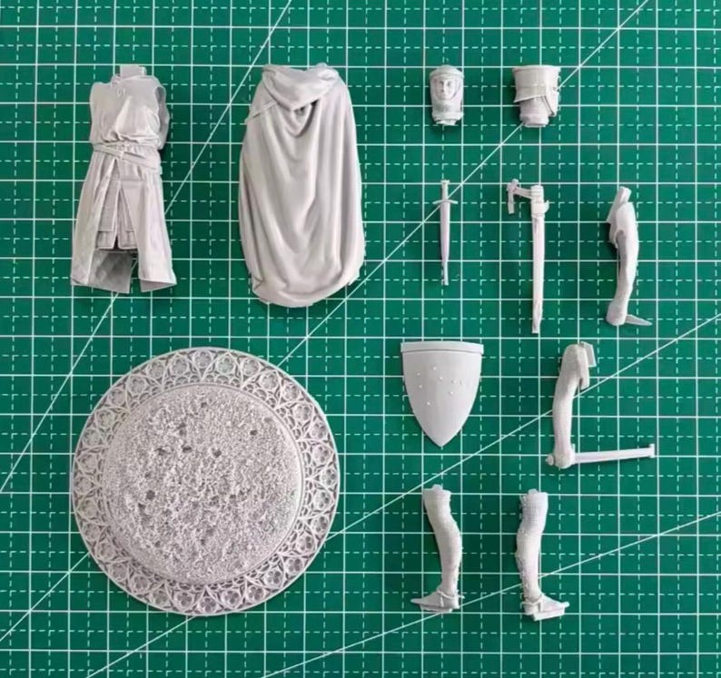May include: A light gray resin model kit consisting of a torso, a hooded cloak, a head, a shield, a sword, and various limb components. The set includes a circular base with a textured surface and an intricate, decorative border pattern.
