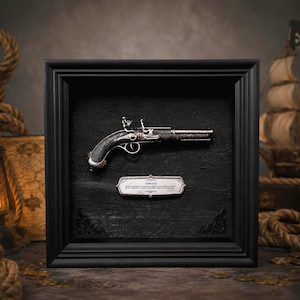 Può includere: Una cornice a cassetta nera contenente una replica di pistola a pietra focaia argentata montata su uno sfondo nero testurizzato. Sotto la pistola si trova una targhetta argentata con il testo FLINTLOCK e informazioni storiche descrittive.