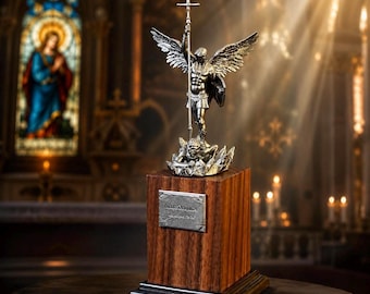 Könnte beinhalten: Silberne Statue eines Engels mit ausgebreiteten Flügeln, der ein Kreuz und einen Schild hält, auf einem Holzsockel. Die Statue steht vor einem verschwommenen Hintergrund eines Kircheninneren mit Buntglas und Kerzen. Eine Plakette befindet sich auf dem Holzsockel.