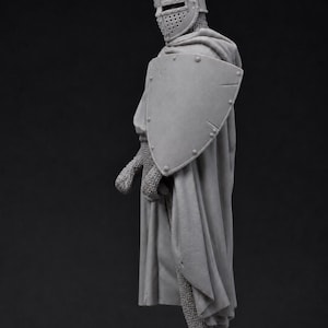 May include: A grey unpainted miniature figurine of a medieval knight standing in profile. The knight wears a flat-topped helmet with a visor, chainmail armor, and a long flowing cloak. A large triangular shield is held against the left side.