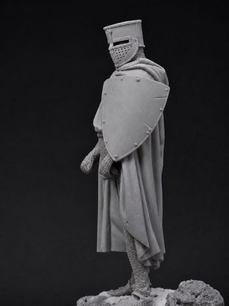 May include: A grey unpainted miniature figurine of a medieval knight standing in profile. The knight wears a flat-topped helmet with a visor, chainmail armor, and a long flowing cloak. A large triangular shield is held against the left side.