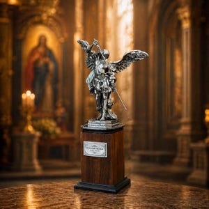 Peut inclure: Une statue argentée détaillée d'un ange aux ailes déployées, tenant une épée, sur un socle en bois foncé. Le socle comporte une petite plaque avec du texte. L'arrière-plan est un intérieur d'église flou avec un éclairage chaleureux.