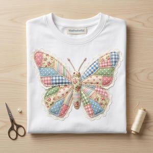 Puede incluir: Sudadera blanca con un diseño de mariposa de patchwork. La mariposa presenta varias telas estampadas en rosa, azul, verde y blanco, con un ribete de encaje. La marca "bluebasketful" es visible.