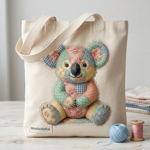 May include: A cream canvas tote bag with a patchwork koala bear design. The koala is made from pastel fabrics with floral and gingham patterns. The bag has cream handles and the text "bluebasketful" is visible on the bottom.