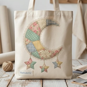 May include: Cream-colored tote bag featuring a patchwork crescent moon design with floral and gingham patterns in pink, blue, and yellow. Three star-shaped charms dangle below the moon. The text "bluebasketful" is printed on a small label.