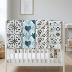 May include: Four baby blankets with different patterns are displayed on a white crib. The patterns include snowflakes, mountains, paw prints, and polar bears. The blankets are in shades of blue, gray, and white.