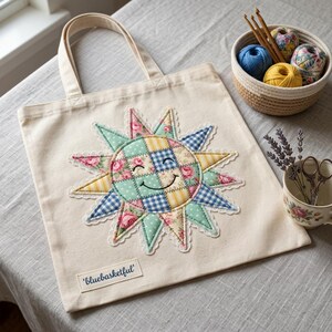May include: Cream-colored tote bag with a large, appliqued sun design. The sun features a patchwork of floral, gingham, and polka dot fabrics in pastel colors. The sun has a smiling face and is surrounded by lace trim. The bag has two cream-colored handles and a label that reads 'bluebasketful'.