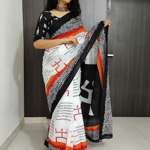 May include: A white saree with a black border and a black and white patterned blouse. The saree features red and orange stripes and a repeating pattern of symbols and text. The blouse has black sleeves with a white pattern.