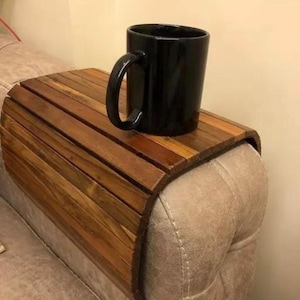 Puede incluir: Una bandeja de brazo de sofá de madera con acabado marrón oscuro, diseñada para descansar sobre un brazo de sofá. Una taza negra está sobre la bandeja. La bandeja está hecha de listones de madera y tiene un diseño curvo.