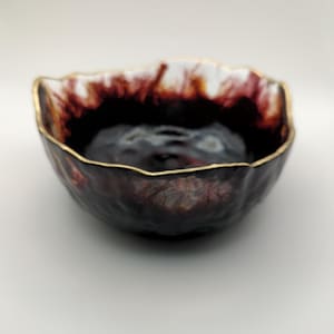 Może przedstawiać: Ręcznie wykonana ceramiczna miska z ciemnym, teksturowanym zewnętrzem i złotym obramowaniem. Wnętrze przechodzi od głębokiego bordowego do jaśniejszego, prawie białego koloru w pobliżu krawędzi. Miska ma organiczny, nieregularny kształt.