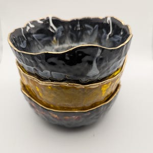 May include: A stack of three decorative bowls with irregular edges and a metallic gold rim. The top bowl is black with white and gray marbling, the middle bowl is gold, and the bottom bowl is dark brown.