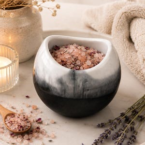 May include: A rounded ceramic bowl with a white top and black bottom gradient finish, filled with pink bath salts mixed with dried flower petals. A small wooden spoon rests nearby, also containing the pink salt and petal mixture.