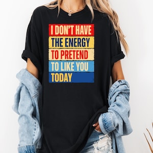 May include: A black short-sleeved t-shirt featuring a graphic print with the text I DON'T HAVE THE ENERGY TO PRETEND TO LIKE YOU TODAY. The text is arranged in five horizontal, distressed-style rectangular blocks coloured red, yellow, and blue.