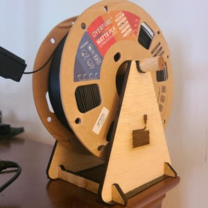 Op de afbeelding: Een spoel zwart Overture Matte PLA 3D-printerfilament op een houten standaard. De spoel heeft een kartonnen label met het Overture-logo en productinformatie. De standaard is gemaakt van lichtgekleurd hout.