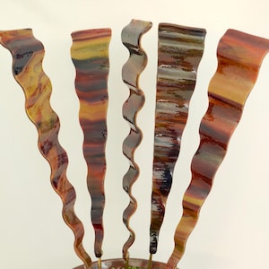 May include: Abstract metal sculpture featuring five vertical, wavy forms in shades of orange, red, yellow, and gray. The sculpture has a textured, painted appearance and is mounted on a circular base.