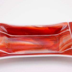 May include: A rectangular, decorative glass dish with a vibrant red and orange color scheme. The dish features a curved design with three distinct sections, bordered by a white edge. The glass has a marbled effect.