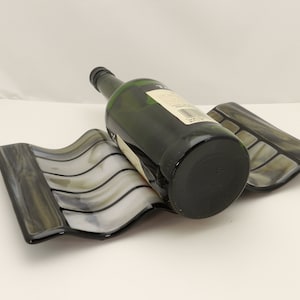 May include: A decorative wine bottle holder with a wavy design in shades of gray, black, and white. A green wine bottle rests on the holder. The holder is made of glass and has a unique, artistic appearance.