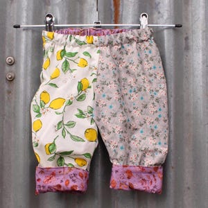 May include: Baby pants with a split design. One side features a white background with yellow lemons and green leaves. The other side has a gray floral pattern. The cuffs and waistband are purple with a floral print.