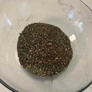 May include: A clear glass bowl filled with a mound of dried herbs. The herbs are a mix of green and brown, with a slightly coarse texture. The bowl is set against a white marble surface.