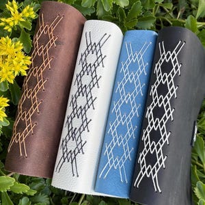May include: Four leather wrist cuffs in brown, white, blue, and black, each with a unique geometric stitched design. The cuffs are arranged on a bed of green foliage with yellow flowers.