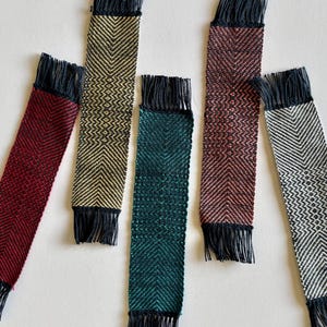 May include: Six handmade woven bookmarks in various colors: burgundy, gold, teal, and red. Each bookmark features a chevron pattern and black fringe at both ends. The bookmarks are arranged on a white surface.