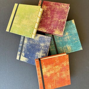 May include: A collection of small, square journals with textured covers in various colors: green, red, blue, teal, and orange. Each journal has a black elastic band closure and is hand-stitched with visible thread. The covers have a distressed, aged appearance.