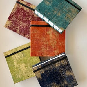 May include: A collection of handmade journals in various colors, including teal, orange, green, burgundy, and navy. Each journal features a textured, distressed design with gold accents, a black elastic band, and stitched binding. The journals are square-shaped.