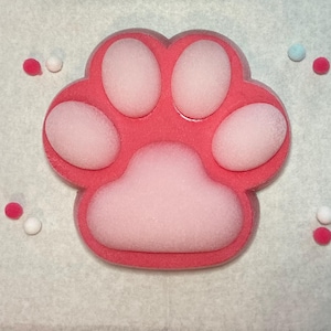 Puede incluir: Un objeto rosa en forma de huella de pata con un centro rosa más claro y detalles blancos. Pequeñas decoraciones redondas y coloridas están dispersas alrededor de la huella. El artículo tiene un aspecto suave y texturizado.