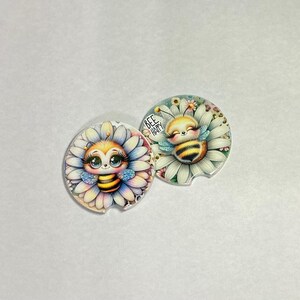 May include: Two round car coasters with a floral and bee theme. One coaster features a bee in a flower with the text "BEE HAPPY". The other coaster has a bee in a flower. The coasters have a white background.