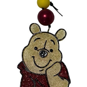 May include: A Winnie the Pooh character ornament featuring a glittery yellow bear wearing a red shirt. The ornament is suspended by a black string adorned with three spherical beads in metallic gold, bright yellow, and a deep, glossy red finish.